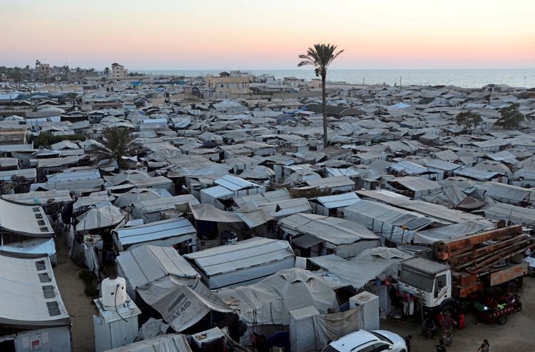 Weitläufiges Zeltlager mit dicht stehenden Zelten in Khan Younis, südlicher Gazastreifen, bei Sonnenuntergang. Im Vordergrund sind ein Lastwagen und Menschen zu sehen. Im Hintergrund das Meer und Gebäude.