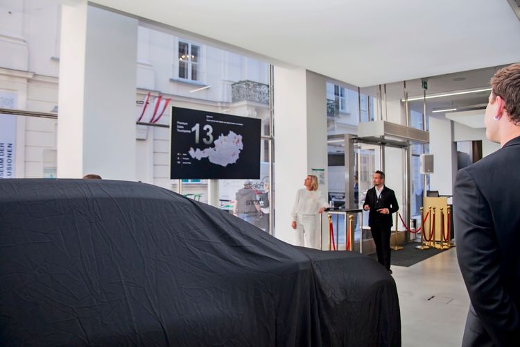 Eine Veranstaltung im Polestar-Space in Wien. Ein abgedecktes Fahrzeug, vermutlich der Polestar 4 SUV, steht im Vordergrund. Im Hintergrund präsentieren zwei Personen vor einer Leinwand mit einer Österreich-Karte und der Zahl „13“.