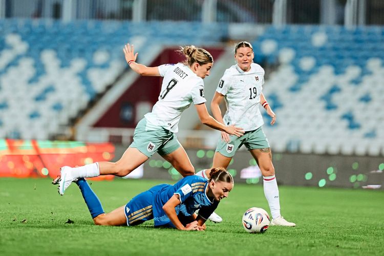 Zwei Fußballspielerinnen des österreichischen Teams in weißen Trikots und grauen Shorts kämpfen um den Ball gegen eine Spielerin des gegnerischen Teams in blauem Trikot, die am Boden liegt. Im Hintergrund ist ein teilweises leerstehendes Stadion zu sehen.