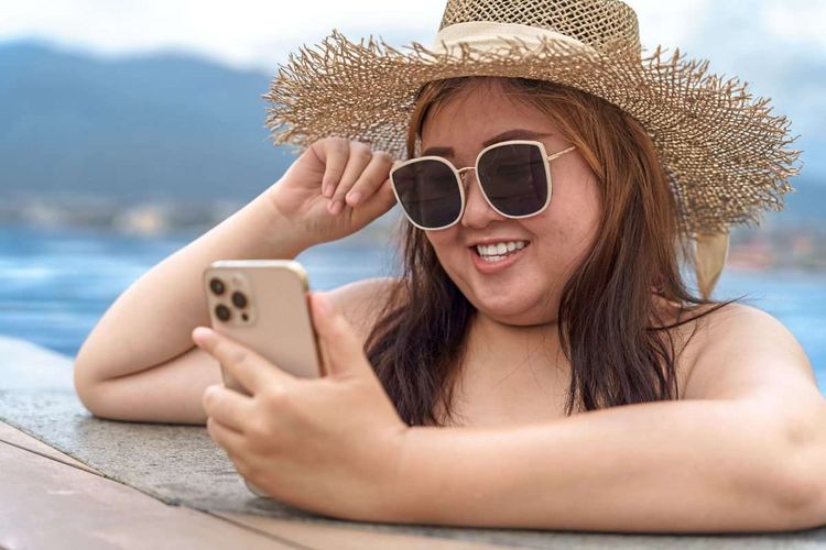 Eine junge, etwas dicke Frau in Pool stützt sich auf den Poolrand und blickt auf ihr Smartphone, sie trägt Sonnenbrille und Sonnenhund.