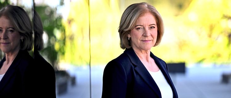 Eine Person mit schulterlangem, blondem Haar steht vor einer Glasfassade, die ihre Spiegelung zeigt. Die Person trägt einen dunklen Blazer und ein weißes Oberteil. Der Hintergrund ist mit unscharfen grünen und blühenden Elementen gefüllt. (Gesicht unkenntlich gemacht).