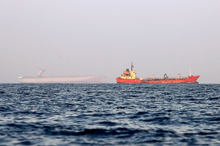 Zwei Handelsschiffe sind auf dem Meer vor Anker zu sehen, eines im Vordergrund in orange-roter Farbe, das andere weiter im Hintergrund und weniger deutlich sichtbar. Der Himmel ist neblig, und das Meer ist ruhig. Die Schiffe befinden sich zwischen den iranischen Inseln Qeshm und Larak in der Straße von Hormus.