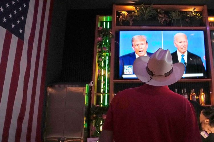 Donald Trump (links) und Joe Biden (rechts) auf einem Fernsehbildschirm. Davor ein Mann mit Cowboyhut von hinten, ganz links eine US-Flagge.