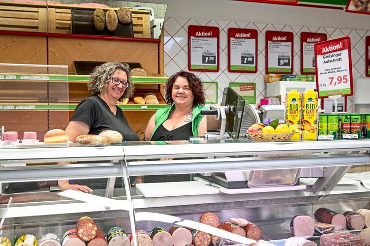 Zwei Frauen stehen lächelnd hinter einer Fleisch- und Wursttheke in einem Lebensmittelgeschäft. Im Vordergrund sind verschiedene Wurstsorten und Lebensmittel zu sehen. Im Hintergrund befinden sich Regale mit Brot und Aktionshinweise.