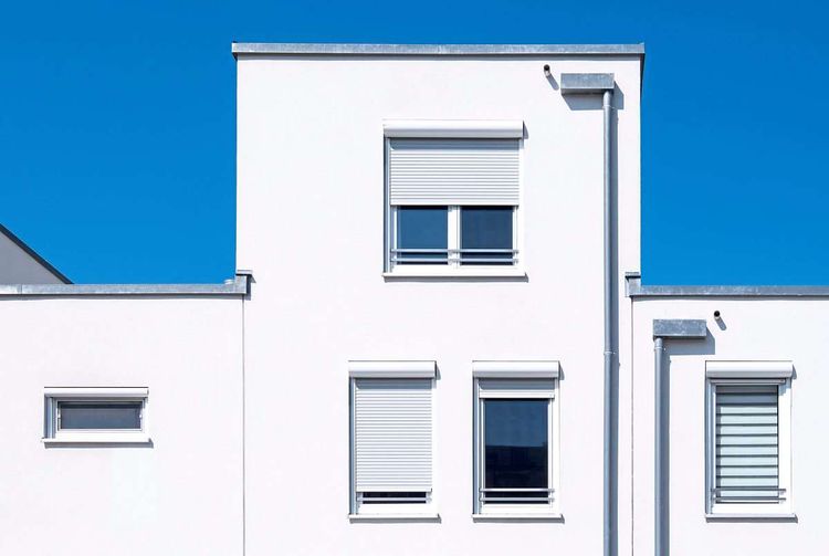 Ein weißer Neubau mit Rollläden, die teilweise geöffnet und geschlossen sind; dahinter strahlend blauer Himmel.