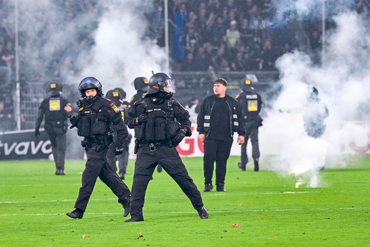 Polizisten in Schutzkleidung sichern ein Fußballfeld, während Rauch und Pyrotechnik zu sehen sind. Im Hintergrund stehen weitere Personen und Sicherheitskräfte. Die Szene deutet auf erhöhte Polizeipräsenz und Sicherheitsmaßnahmen bei einem Hochrisikospiel hin.