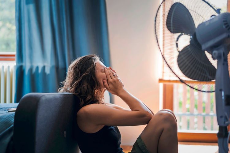 Junge Frau sitzt erschöpft auf dem Boden vor einem Ventilator und bedeckt ihr Gesicht mit den Händen, vermutlich wegen der Hitze.