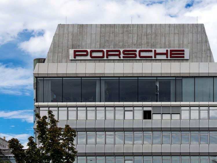 Das Bild zeigt ein modernes Gebäude mit dem Schriftzug „PORSCHE“ in roten Buchstaben auf einem weißen Hintergrund an der Spitze. Es hat große Fensterfronten im oberen Bereich und ist von blauem Himmel mit vereinzelten Wolken umgeben. Im Vordergrund sind Baumspitzen sichtbar.