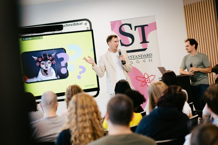 Eine Präsentation bei einer Veranstaltung: Eine Person in einem cremefarbenen Anzug spricht mit Mikrofon vor einem Publikum, im Hintergrund ein Bildschirm mit einem Bild von einer Katze mit Lockenwicklern und Fragezeichen sowie ein Banner mit der Aufschrift 