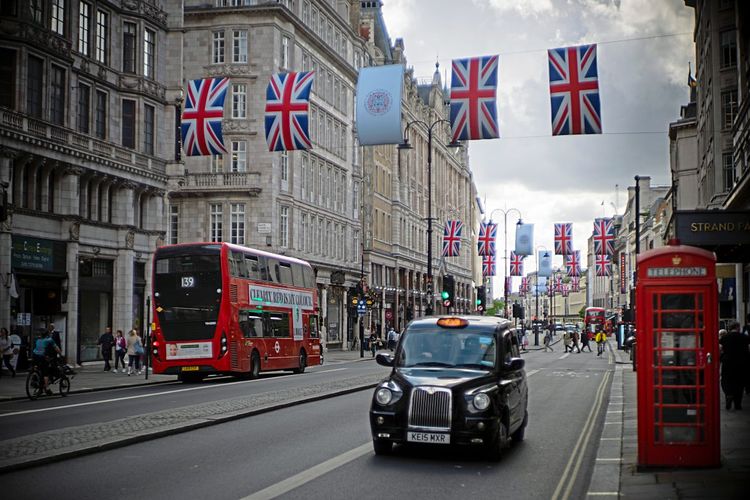 Rechts fährt ein schwarzes Taxi, links ein Doppeldeckerbus. An einer Leine über den Fahrzeugen hängen vier britische Flaggen.