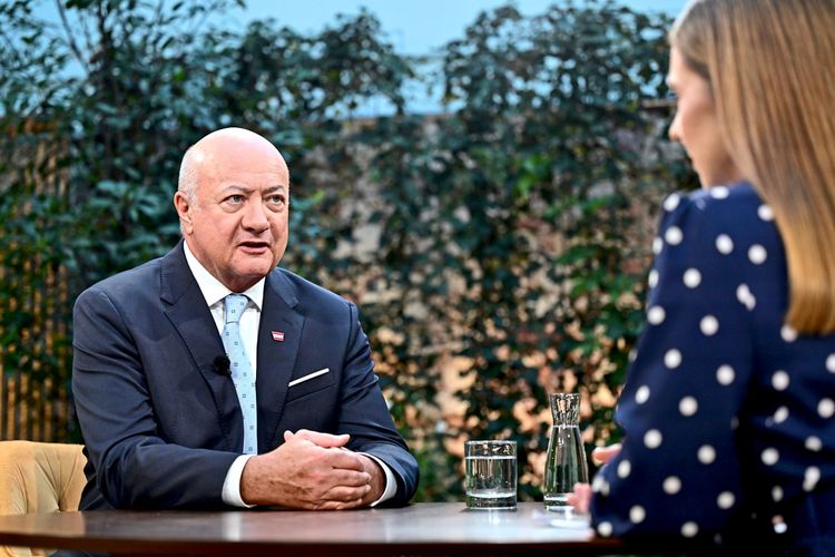 Ein Mann im Anzug spricht in einem Interview mit einer Frau, die einen dunkelblauen, gepunkteten Blazer trägt. Beide sitzen an einem Tisch mit Gläsern und einer Wasserkaraffe. Im Hintergrund ist grüne Vegetation zu sehen.