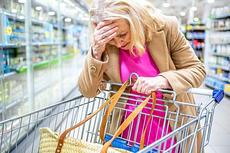 Eine ältere, sorgenvoll blickende Frau stützt sich im Supermarkt auf einen leeren Einkaufswagen.