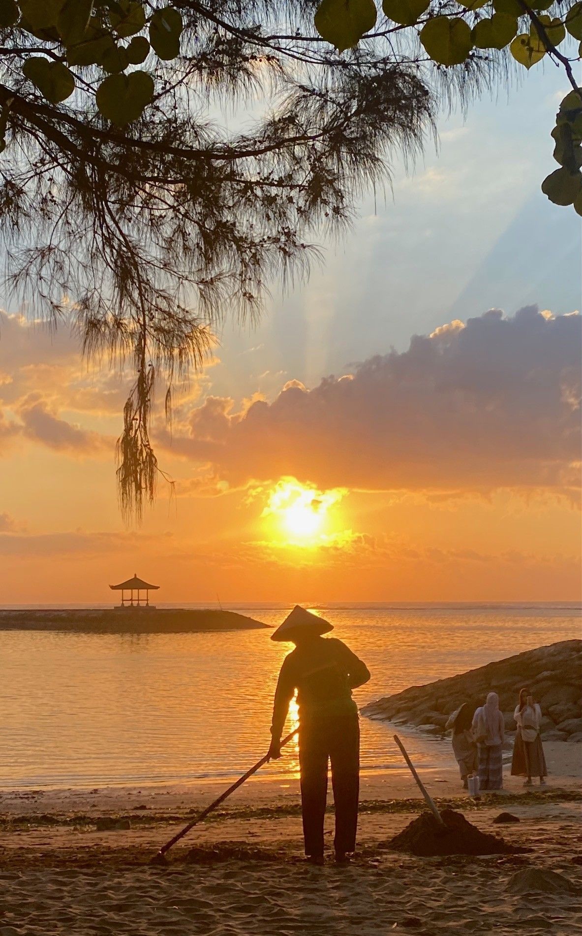 Ein Mann mit Rechen auf einem Sandstrand bei Sonnenaufgang, im Hintergrund Menschen und das Meer