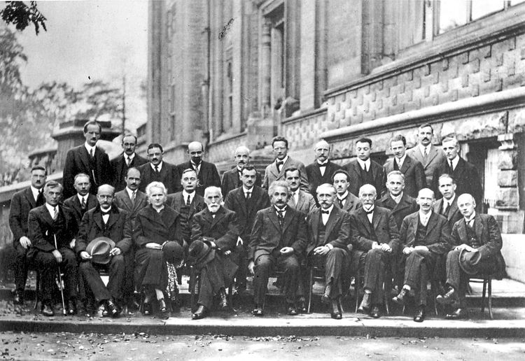 Schwarz-Weiß-Fotografie einer großen Gruppe von Männern und Frauen in formeller Kleidung, die vor einem historischen Gebäude posieren. Die Personen sitzen und stehen auf unterschiedlichen Ebenen, einige halten Hüte in den Händen. Die Aufnahme stammt vom fünften Conseil Solvay für Physik im Jahr 1927 in Brüssel, einer renommierten wissenschaftlichen Konferenz, die viele bedeutende Physiker der Zeit zusammenbrachte.