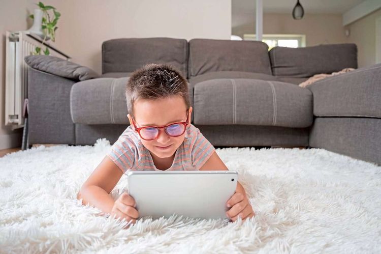 Ein kleiner Bub mit Brille liegt mit einem Tablet in den Händen auf einem flauschigen Teppich auf einem Wohnzimmerboden