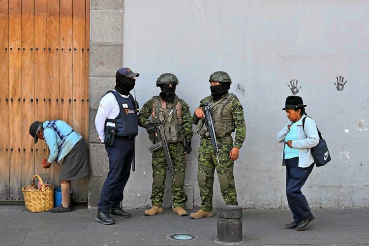 Zwei bewaffnete Soldaten in Tarnuniform und ein maskierter Sicherheitsbeamter stehen auf einer Straße in Quito, Ecuador. Links kniet eine ältere Frau neben einem Korb, und rechts geht eine weitere ältere Frau vorbei. Im Hintergrund sind eine Holztür und eine graue Wand mit Handabdrücken zu sehen.