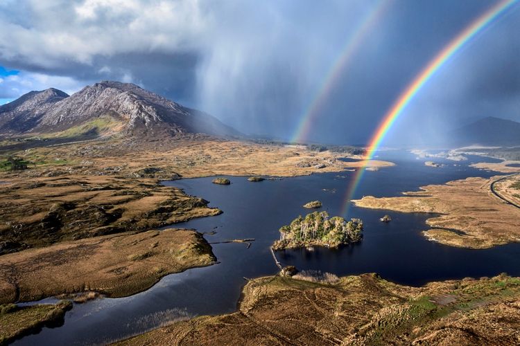 Regenbogen über dem Derryclare Lough, einem Süßwassersee in Connemara, Irland. Im Vordergrund eine kleine, mit Bäumen bewachsene Insel im See, umgeben von weitem Moorland. Im Hintergrund ragen majestätische Berge unter dramatischem Himmel auf. Ein zweiter, schwächerer Regenbogen ist ebenfalls sichtbar.