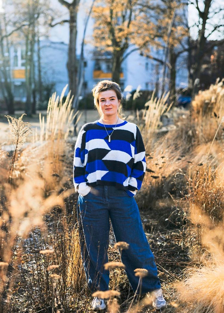 Eine Frau steht auf einer Wiese mit vertrocknetem Gras und Pflanzen. Sie trägt einen farbenfrohen Pullover in Blau, Weiß und Schwarz mit geometrischem Muster sowie blaue Jeans. Im Hintergrund sind verschwommen Bäume und ein Gebäude zu sehen. Die Szene ist in warmem Sonnenlicht aufgenommen.