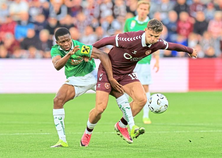Zwei Fußballspieler kämpfen um den Ball während eines Spiels. Der Spieler in bordeauxrot versucht, den Ball zu schützen, während der Spieler in grün-weiß versucht, ihn zu erobern. Im Hintergrund sind weitere Spieler und verschwommene Zuschauer zu sehen.