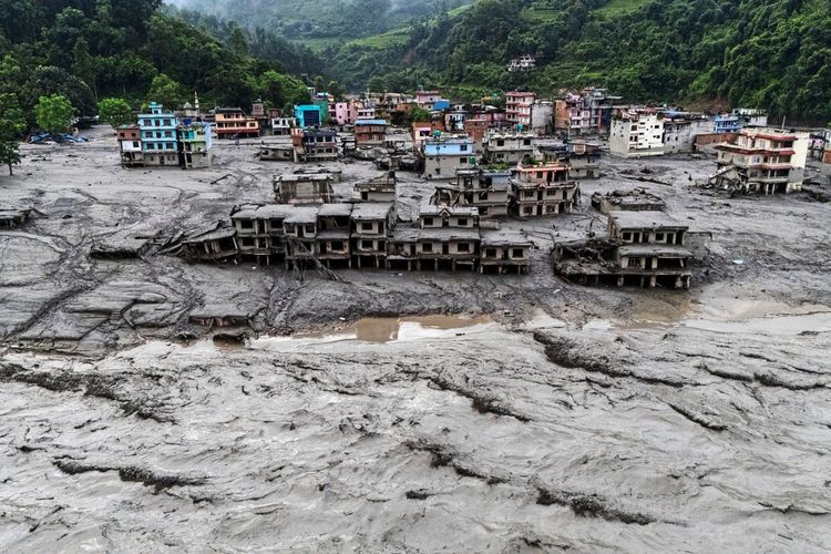 Luftaufnahme einer Flutkatastrophe in Devighat, Nuwakot, Nepal. Gebäude sind schwer beschädigt und von Schlamm und Wasser bedeckt, nachdem tödliche Sturzfluten und Erdrutsche die Region heimgesucht haben. Im Hintergrund sind grüne Hügel zu sehen.