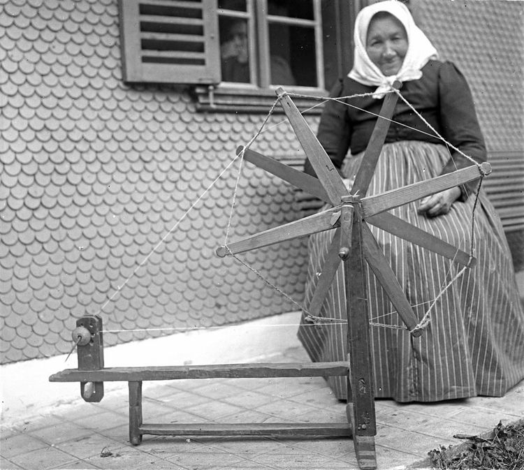 Eine Bäuerin im Montafon sitzt an einem Baumwollrad und spinnt.