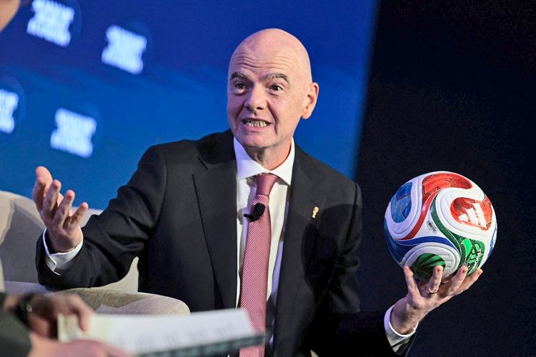 Ein Mann in einem Anzug und mit einer roten Krawatte hält einen Fußball in der Hand und gestikuliert, während er spricht. Der Hintergrund zeigt eine Bühne mit verschwommenem Text.
