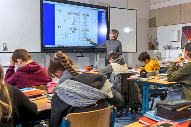 Eine Lehrerin unterrichtet Mathematik in einer 7. Klasse in einem Klassenzimmer mit einem interaktiven Whiteboard. Die Schülerinnen und Schüler sitzen an ihren Tischen, schreiben mit und arbeiten konzentriert. Auf dem Whiteboard ist eine Aufgabe zu Gewicht, Preis, Wegstrecke und Zeit zu sehen. Im Raum sind Schultaschen und Materialien verteilt.