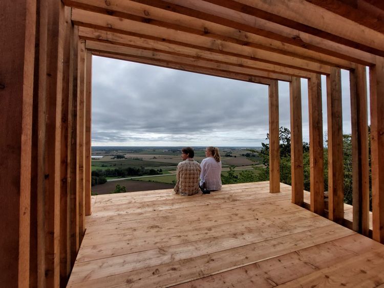 Zwei Menschen sitzen auf einem Holzgerüst und blicken auf die Landschaft.