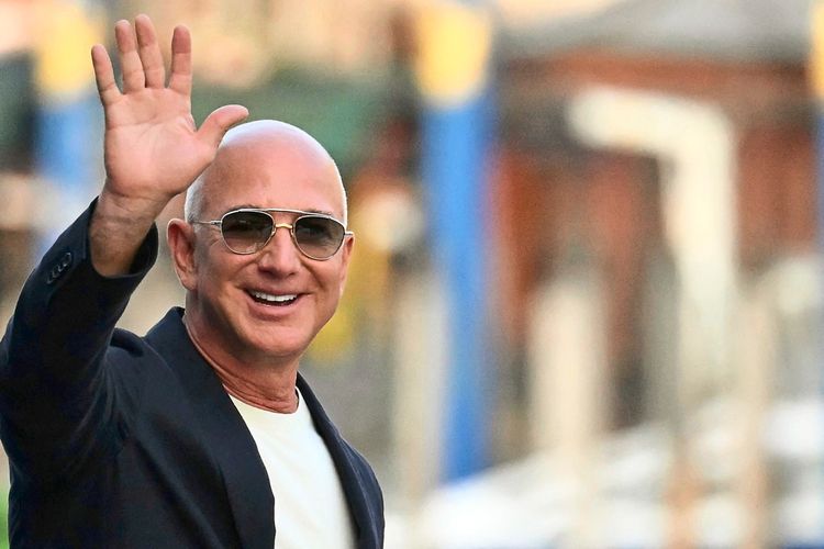 Ein Mann mit Glatze und Sonnenbrille winkt fröhlich in die Kamera. Er trägt ein schwarzes Jacket über einem weißen Hemd, der Hintergrund ist unscharf.