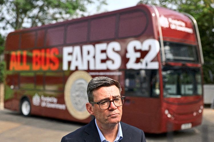 Andy Burnhham steht mit Brille vor einem roten Doppeldeckerbus mit der Aufschrift „ALL BUS FARES £2“. Im Hintergrund Bäume und der Bus, im Vordergrund der Mann, der seriös blickt.