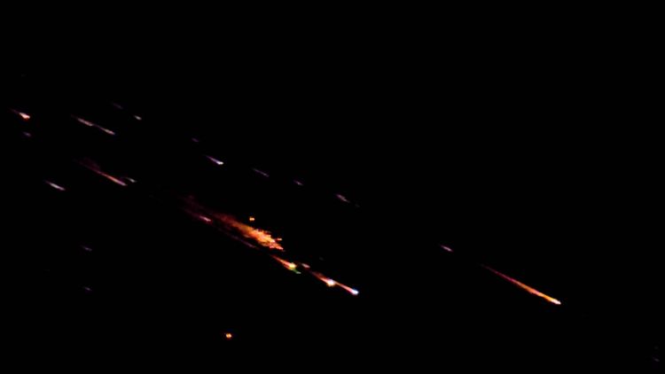 Am Nachthimmel ist ein heller Lichtschweif zu sehen, der durch verglühenden Weltraumschrott entsteht. Die Szenerie ist dunkel mit leuchtenden, orangefarbenen und weißen Lichtspuren, die sich diagonal bewegen.