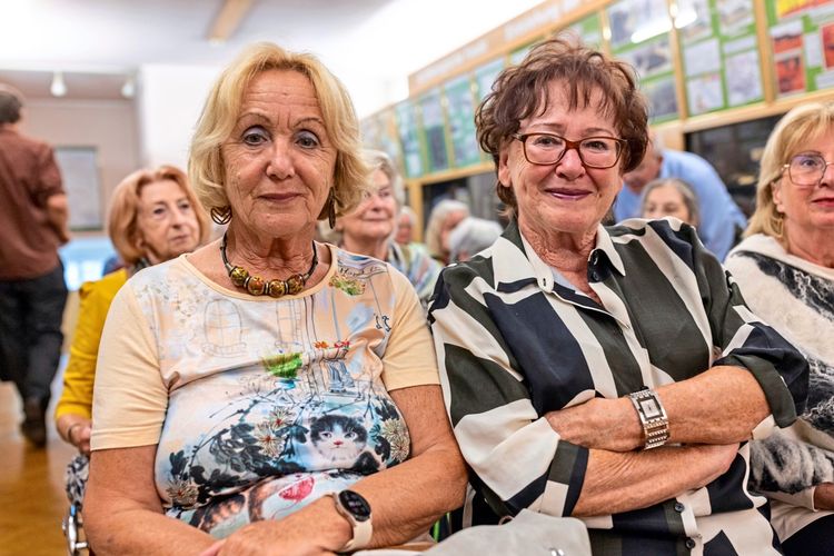 Zwei Frauen sitzen in einem Raum mit anderen Menschen im Hintergrund. Die Frau links trägt ein buntes Shirt mit einem Katzenmotiv, die Frau rechts eine gestreifte Bluse und eine Brille. Beide lächeln leicht in die Kamera. Im Raum sind Ausstellungstafeln an der Wand zu sehen.