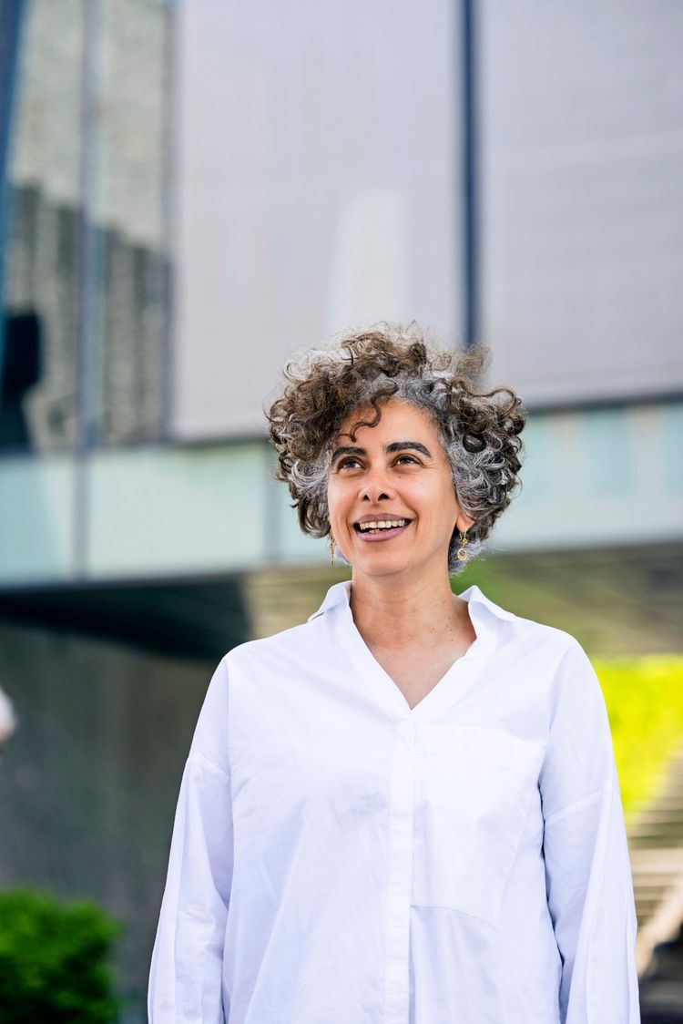 Eine Frau mit lockigem, grauem Haar und einem weißen Hemd lächelt und schaut nach oben. Im Hintergrund sind unscharf moderne Glas- und Betonarchitektur sowie grüne Pflanzen zu sehen.