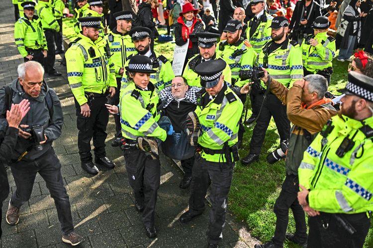 Eine Gruppe von Polizist*innen in leuchtend gelben Uniformjacken trägt einen Demonstranten während einer Protestaktion ab. Der Vorfall findet im Freien statt, umgeben von weiteren Menschen, die zuschauen, fotografieren oder filmen. Im Hintergrund sind weitere Demonstrierende und Gegenstände zu erkennen.