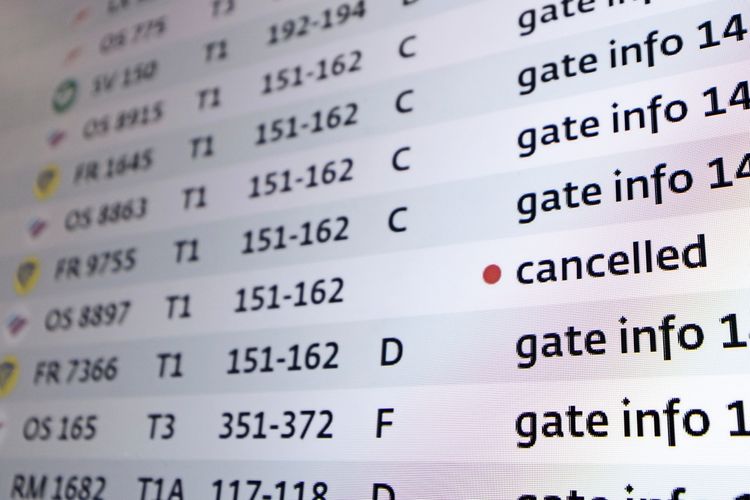 Eine Nahaufnahme einer Informationsanzeige am Flughafen Wien, die Flugdetails zeigt. Eine Zeile ist mit rotem Punkt und dem Hinweis „cancelled“ markiert, was auf einen gestrichenen Flug hinweist. Anderen Zeilen enthalten Informationen zu Gates und Terminalbereichen.