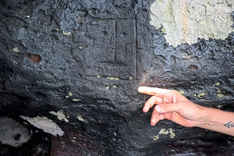 Schwarzer Fels mit hellgrünen Flechten, in den ein rechteckiges Gesicht mit geradem Mund und einer Linie als Nase eingeritzt wurde, auf das eine Hand zeigt.