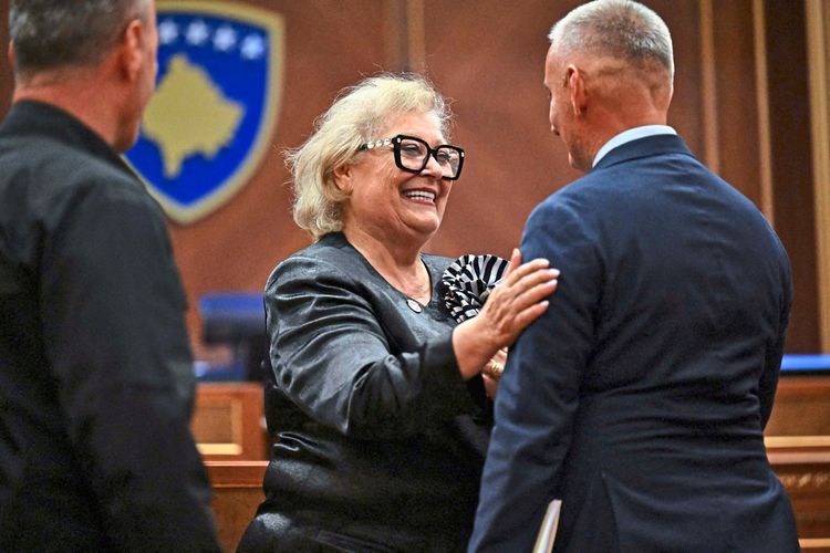 Eine Frau in dunkler Kleidung und Brille, die lächelnd einem Mann gratuliert oder sich mit ihm unterhält. Im Hintergrund ist ein Wappen von Kosovo erkennbar. Die Szene findet in einem offiziellen Raum statt.