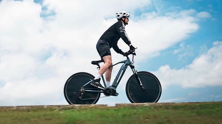 Eine Person fährt ein modernes Fahrrad mit aerodynamischen Scheibenrädern auf einem grasbewachsenen Weg. Sie trägt einen Helm, schwarze Kleidung und weiße Socken. Der Himmel ist blau mit weißen Wolken.