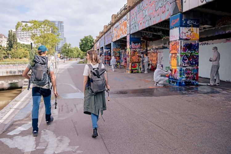 Zwei Menschen gehen mit Angeln den Donaukanal entlang. Rechts von ihnen werden Graffiti gemalt