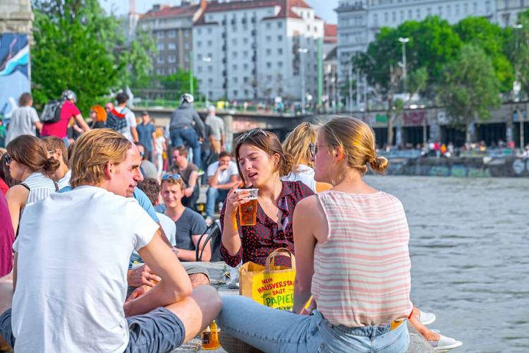 Junge Menschen sitzen am Donaukanal, trinken Bier und unterhalten sich.