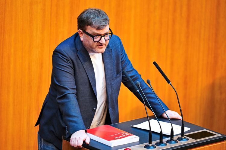 Egy személy áll egy mikrofonokkal ellátott pulpitus mögött. A pulpituson egy piros fedelű könyv és néhány dokumentum található. A háttér fa lambéria.