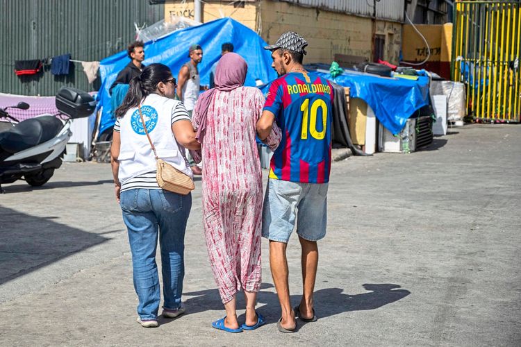 Drei Personen gehen in einem Lagerbereich. Eine Frau in rosa Kleidung und Kopftuch wird von zwei Personen gestützt: links eine Frau mit weißem T-Shirt und Jeans, rechts ein Mann in einem Fußballtrikot mit der Aufschrift „Ronaldinho“. Im Hintergrund sind provisorische Unterkünfte mit blauen Planen, ein Motorrad und weitere Menschen sichtbar.