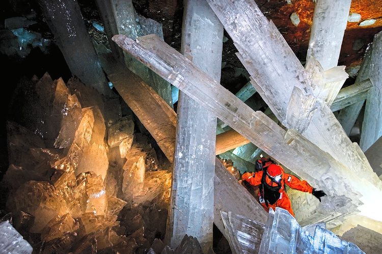 Ein Höhlenforscher in orangefarbenem Anzug geht zwischen meterlangen, weiß-opaken Kristallen hindurch.
