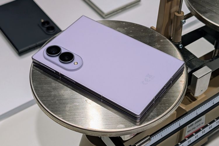 Ein fliederfarbenes, zugeklapptes Falt-Smartphone liegt flach auf der runden Metallschale einer mechanischen Zeigerwaage. Auf der nach oben zeigenden Rückseite befindet sich links oben ein ovales Kameramodul mit zwei großen Kameralinsen und einem kleinen Blitz, während unten rechts dezent gedruckte Prüf- und Entsorgungssymbole zu sehen sind. Im Anschnitt des Bildhintergrunds sind der Messmechanismus der Skala sowie ein weiteres, dunkles Smartphone zu erkennen.