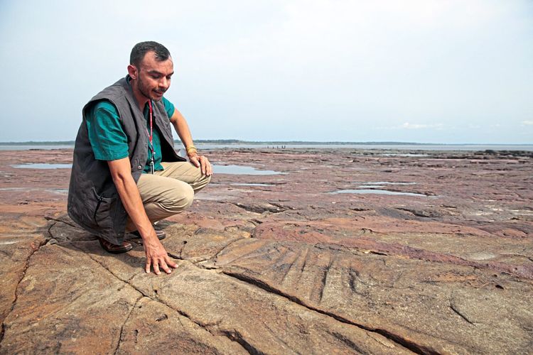Archäologe Jaime de Santana Oliveira geht auf rötlich-gelbem Gestein mit länglichen Einritzungen in die Hocke.