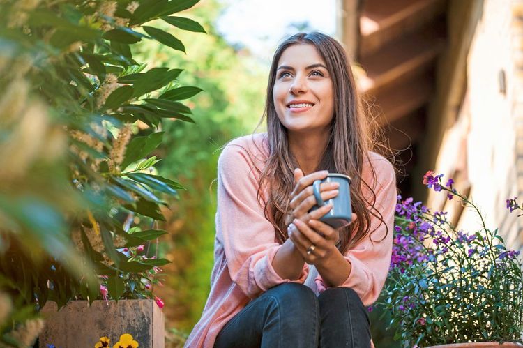Eine Frau mit verklärtem Gesichtsausdruck und Kaffeehäferl in der Hand sitzt entspannt in einem prachtvollen Garten