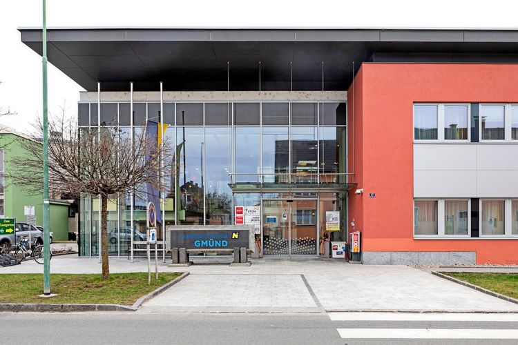 Ansicht des Eingangsbereichs des Landesklinikums Gmünd in Niederösterreich, Österreich. Das moderne Gebäude hat eine Glasfassade, einen roten Seitentrakt und ein flaches Dach. Vor dem Eingang stehen Fahnenmasten und ein Baum. Ein Zebrastreifen führt zum Eingang.