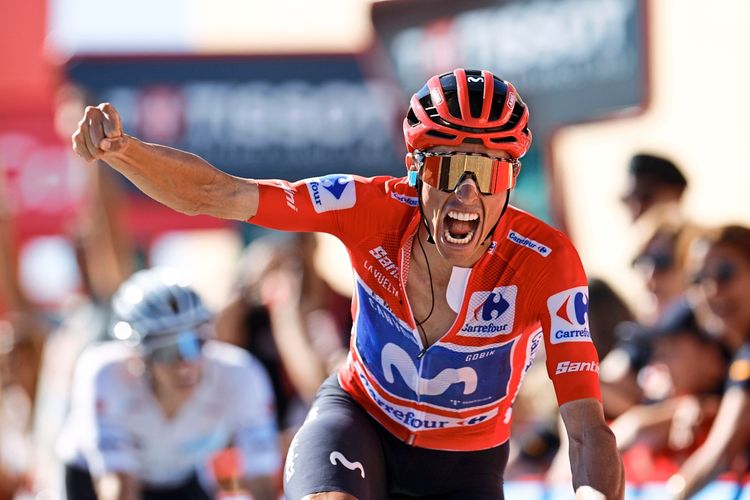 Ein Radfahrer im roten Trikot feiert mit erhobener Faust während einer Etappe der Vuelta a España 2026. Im Hintergrund sind unscharfe Zuschauer und ein weiterer Fahrer zu sehen.