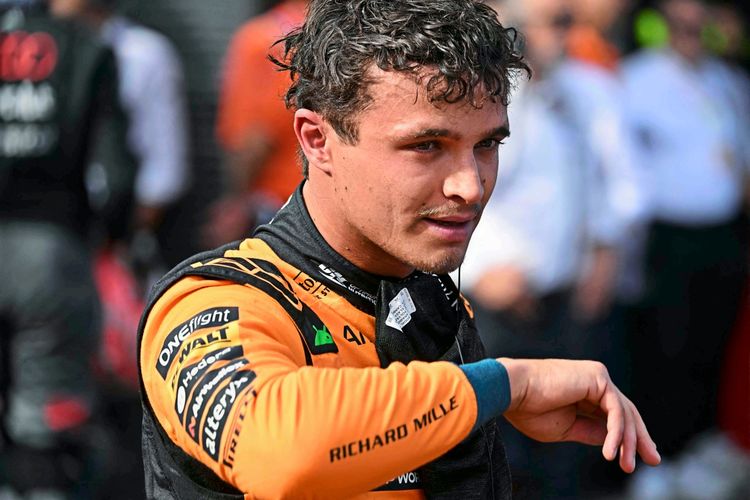 Ein Formel-1-Fahrer im orangefarbenen Rennanzug von McLaren steht nach einer Qualifikationssession auf der Rennstrecke Hungaroring. Im Hintergrund sind unscharfe Personen und eine Rennsportumgebung sichtbar.