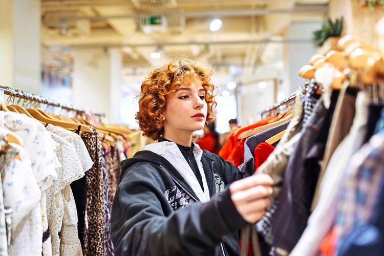 Eine junge Frau mit roten Locken durchsucht Kleidungsstücke in einem Geschäft. Sie betrachtet aufmerksam die Auswahl an herbstlicher Kleidung.